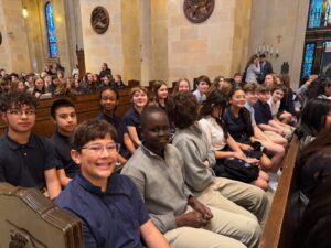 large group of eighth grade students sit in church pews together and smile at camera
