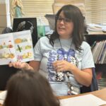 Female teacher facing a classroom of students, reading an open book